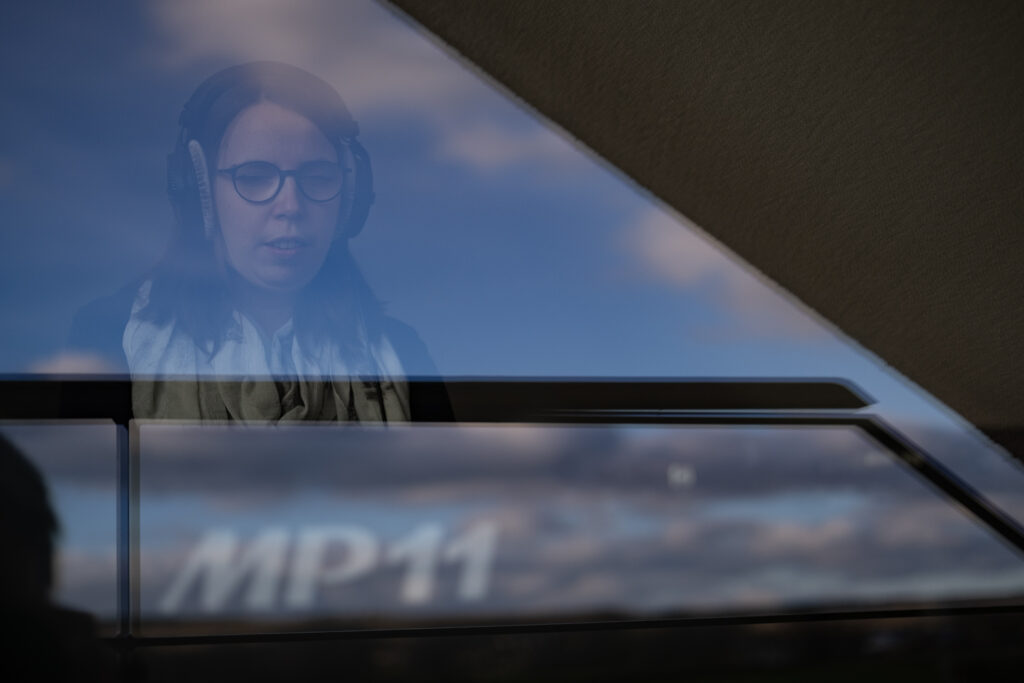 Blick durch ein Fenster, in dem sich blauer Himmel und Schleierwolken spiegeln: Lorena Dürnholz trägt einen Kopfhörer und sitzt mit geschlossenen Augen am Stagepiano