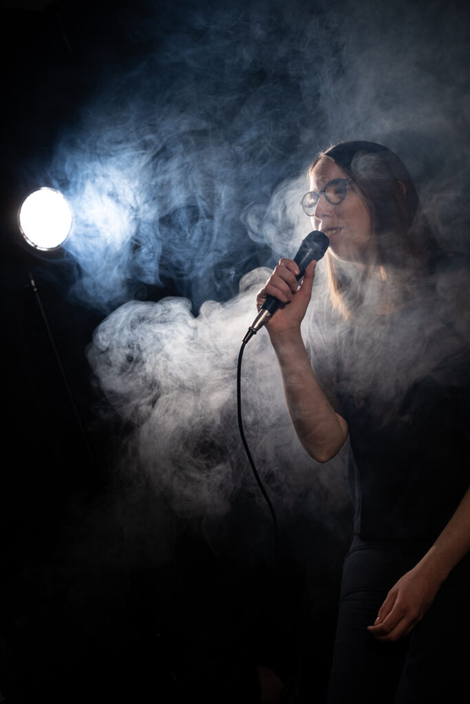 Lorena Dürnholz steht mit einem Mikrofon vor dunklem Hintergrund zwischen Bühnennebel im Scheinwerferlicht und singt