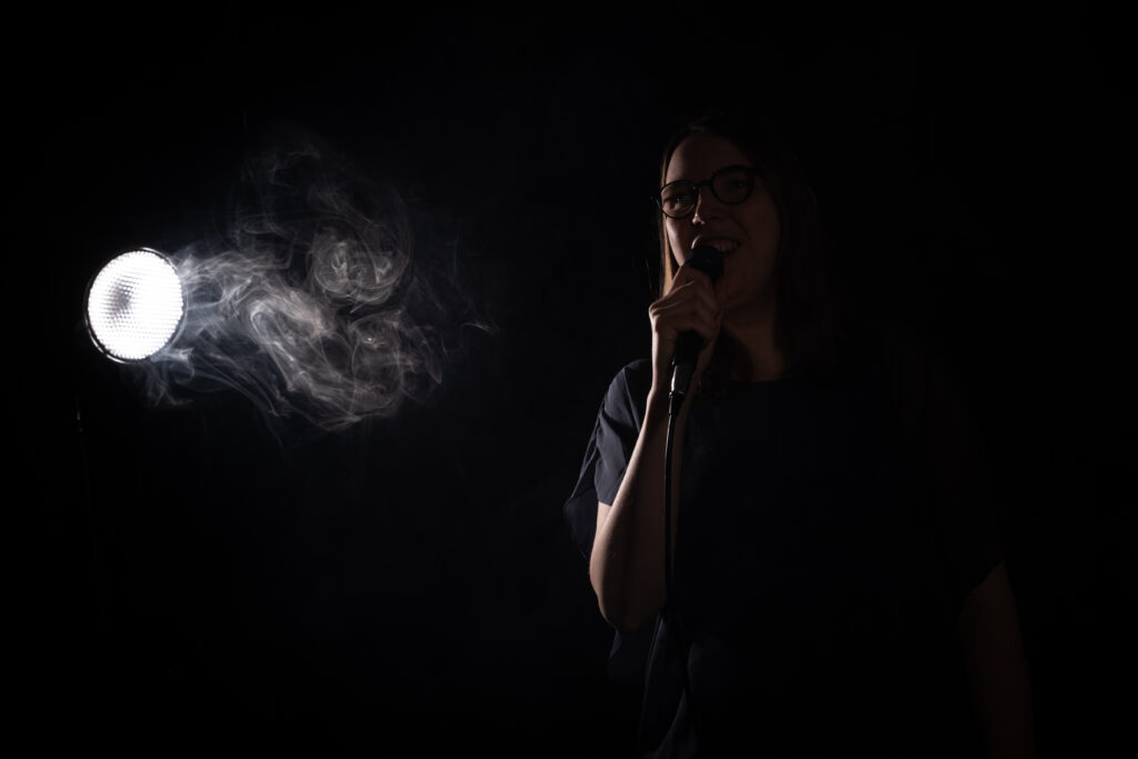 Lorena Dürnholz steht mit einem Mikrofon vor dunklem Hintergrund im Scheinwerferlicht und singt – vor dem Scheinwerfer ist leichter Bühnennebel zu erkennen