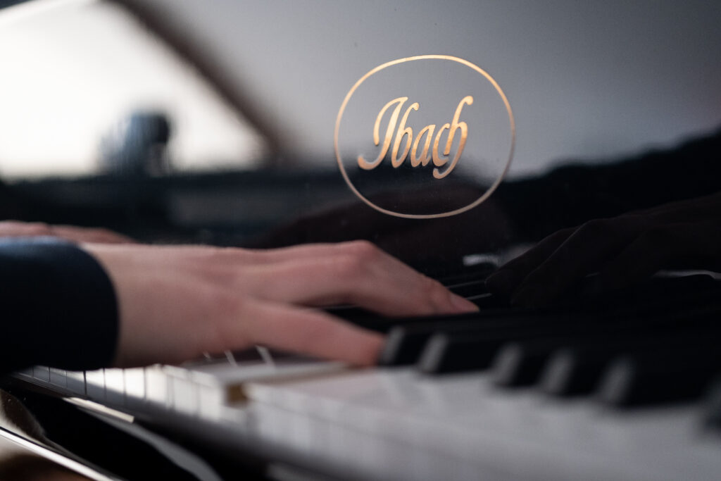 Nahaufnahme von Händen an einem Klavier, auf dem in goldenen Buchstaben der Schriftzug des Herstellers "Ibach" zu lesen ist