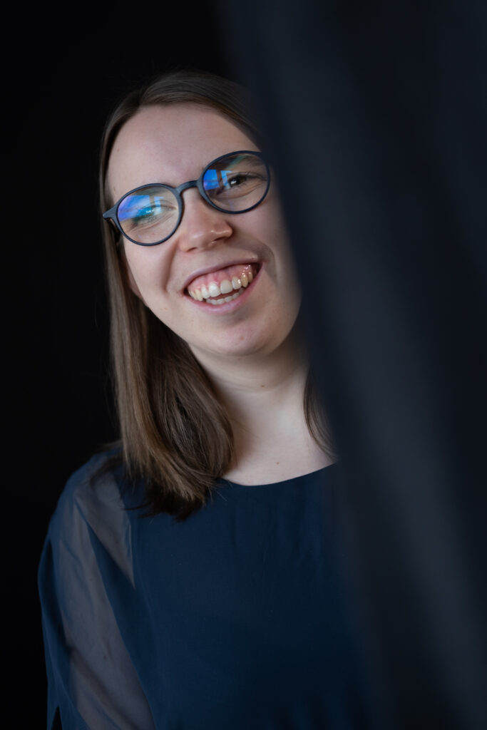 Portraitaufnahme: Lorena Dürnholz, die schulterlange Haare und eine Brille trägt, lächelt in die Kamera