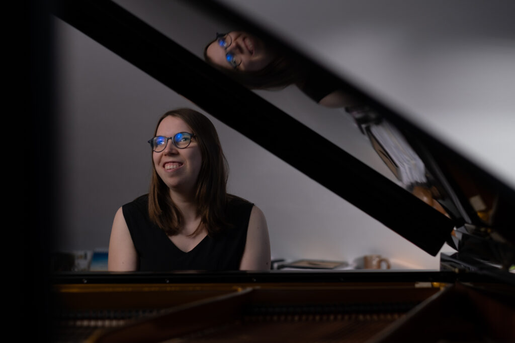 Lorena Dürnholz sitzt lächelnd an einem Konzertflügel und spielt Klavier – im geöffneten Deckel des Instruments spiegelt sich ihr Gesicht