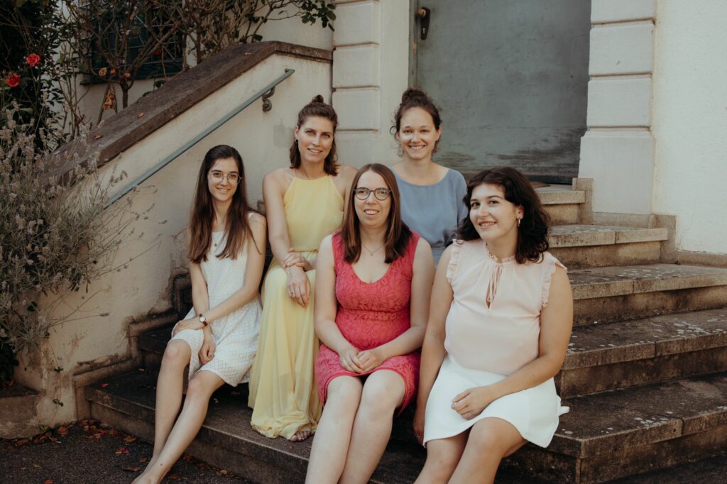 Von Links nach Rechts: Marie, Ariane, Lorena, Tabea und Amelie sitzen auf einer Treppe in Augsburg, in sommerlichen Kleidern und lächeln in die Kamera. 